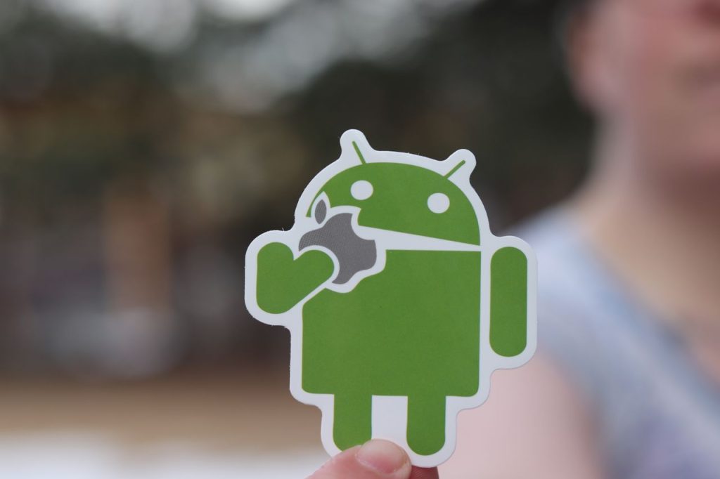 Close-up of an Android sticker, humorously holding an apple logo, with a blurred outdoor background.